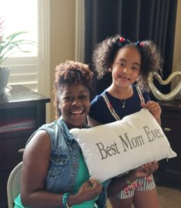 Mom and daughter smile together holding a pillow that says "best mom ever"